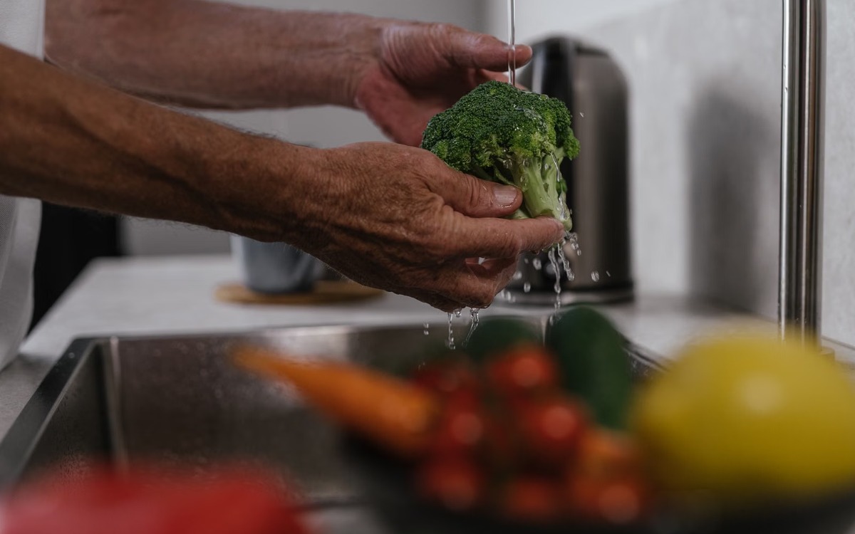 Hands washing fresh broccoli under running water in a kitchen sink