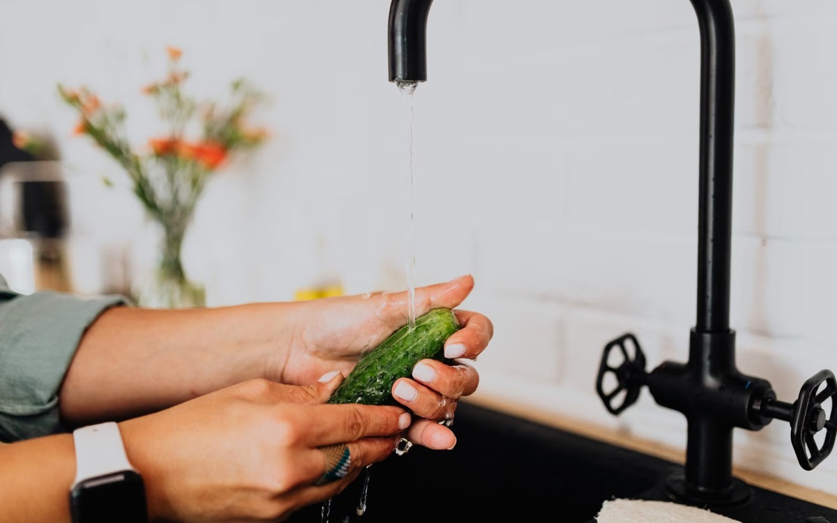 A fresh cucumber being washed under running water in a kitchen sink