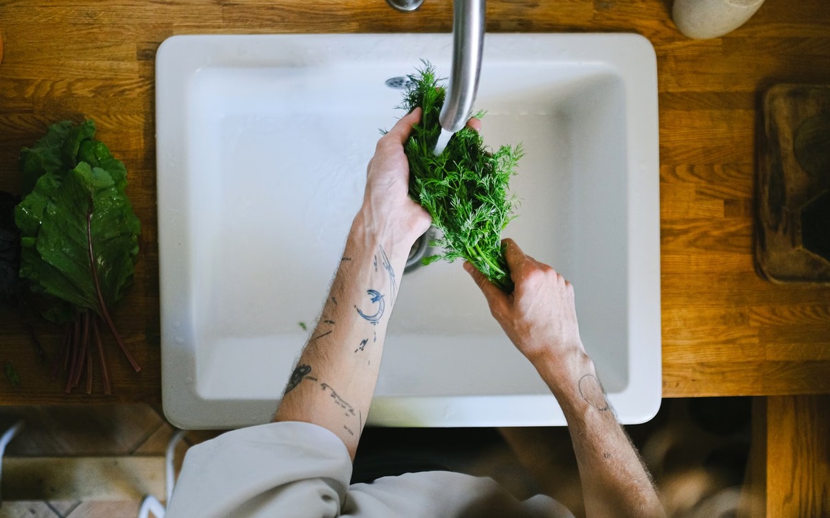 Fresh herbs being rinsed under running water in a kitchen sink